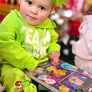 Dahlia is registered to the contest to win money with this photo: child, toddler, green_clothing, book, reading, headband, bow, indoor, cute, holiday_theme, face, eyes, sitting, floor, toy, colorful, expression, person, young_child, playful