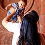 baby, child, adult, family, holding, smiling, happy, portrait, people, hands, hair, clothing, blue_shirt, white_shirt, tattoo, indoor, stone_wall, curly_hair, back_view, interaction