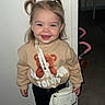 toddler, child, smiling, indoor, pigtails, sweatshirt, teddy_bear, white_purse, black_pants, white_sneakers, floor, wall, doorway, stroller, furniture, happy, cute, person, portrait, fashion