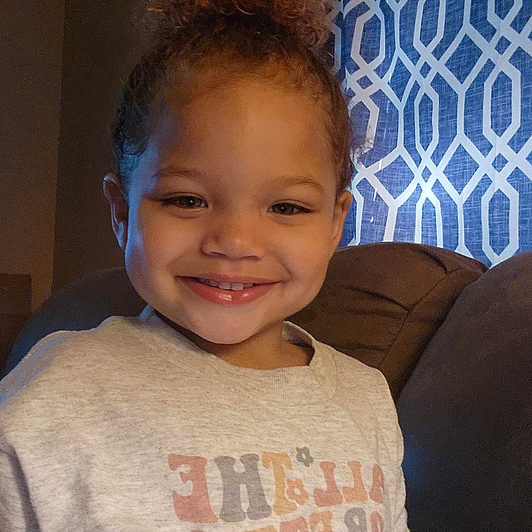 Kahlanie is registered to the contest to win money with this photo: bun, casual_clothing, child, couch, cozy, curly_hair, cute, face, happy, home, indoor, patterned_curtain, person, portrait, relaxed, seated, smiling, sweatshirt, warm_lighting, young_child