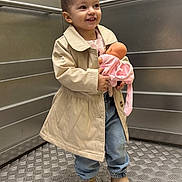 Kiana participe au concours pour gagner de l'argent avec cette photo : beige_coat, boots, casual_clothing, child, cute, doll, elevator, floor_pattern, happy, holding, indoor, jeans, metal_wall, person, play, portrait, smiling, standing, toy, young_child