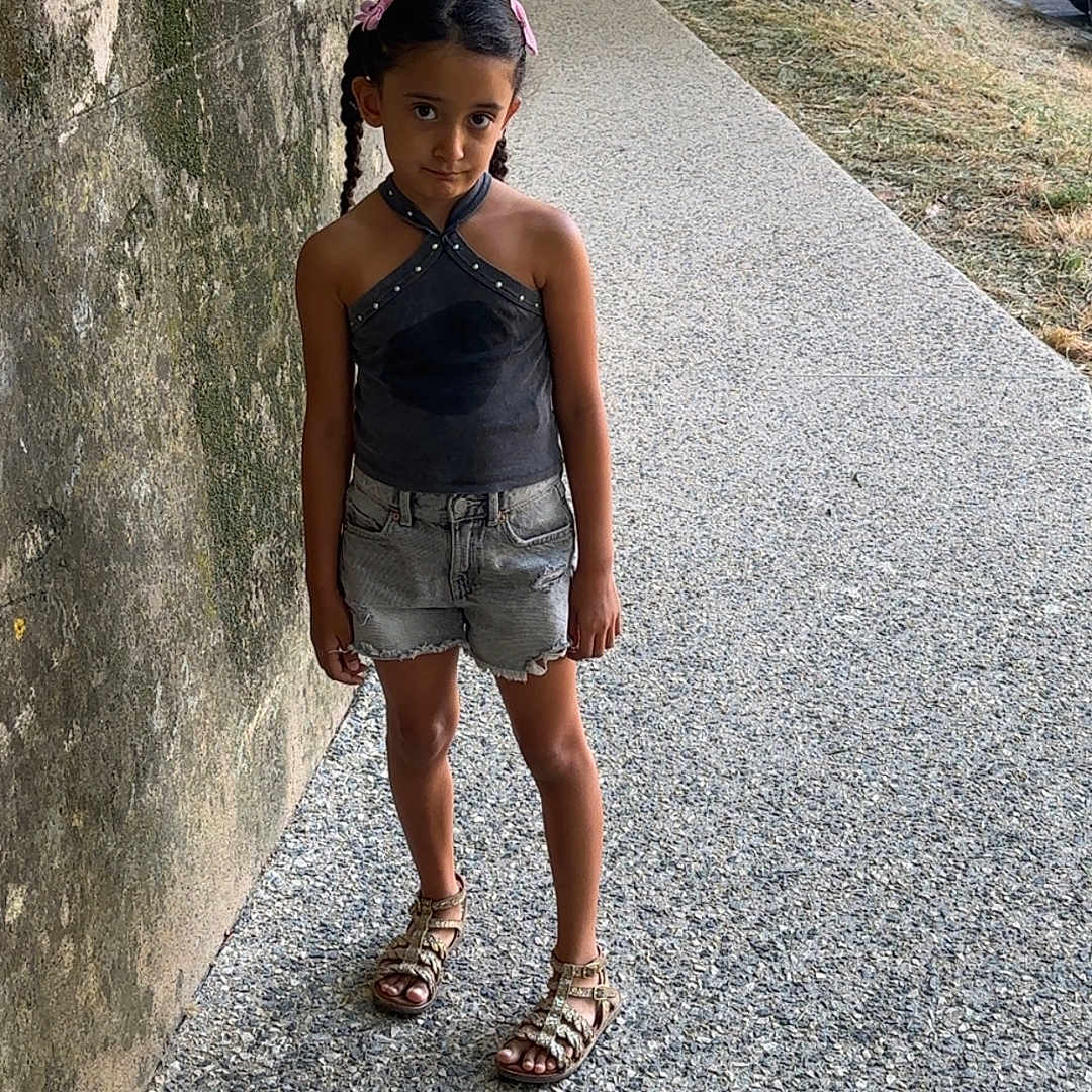 Nelya participe au concours pour gagner de l'argent avec cette photo : black_top, braids, casual_clothing, child, curious_expression, daylight, denim_shorts, girl, mossy_wall, nature, outdoor, parked_cars, person, pink_bows, sandals, sidewalk, standing, summer, trees, urban