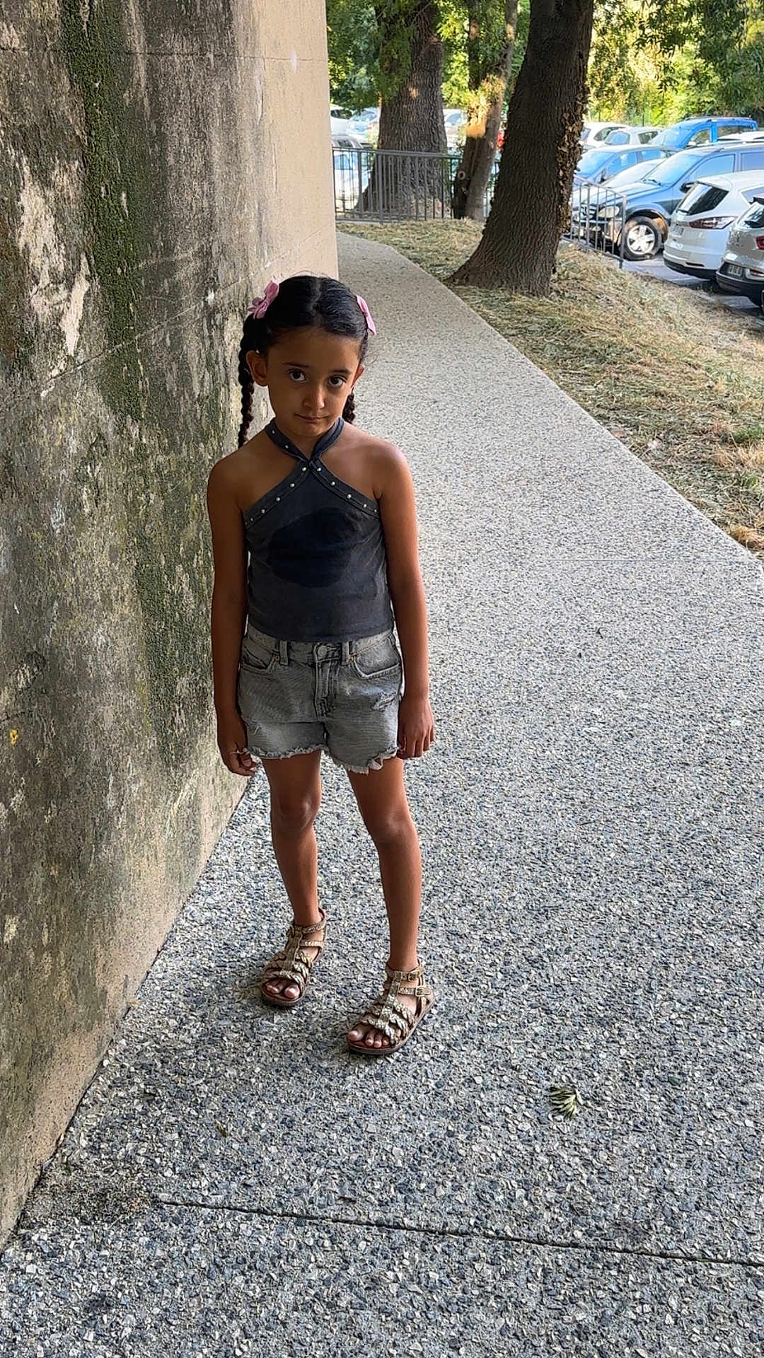 Nelya participe au concours pour gagner de l'argent avec cette photo : black_top, braids, casual_clothing, child, curious_expression, daylight, denim_shorts, girl, mossy_wall, nature, outdoor, parked_cars, person, pink_bows, sandals, sidewalk, standing, summer, trees, urban