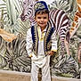 animal_painting, barefoot, blue_vest, boy, child, costume, cute, decor, floor, indoors, leaves, plants, portrait, smile, standing, traditional_clothing, wall_mural, white_pants, wooden_floor, zebra