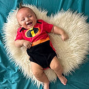 Ramón Jr is registered to the contest to win money with this photo: baby, smiling, infant, red_clothing, black_clothing, incredibles_logo, fluffy_rug, heart_shape, blue_background, lying_down, happy, cute, child, portrait, indoors, skin, bare_feet, short_sleeve, infant_fashion, baby_pose