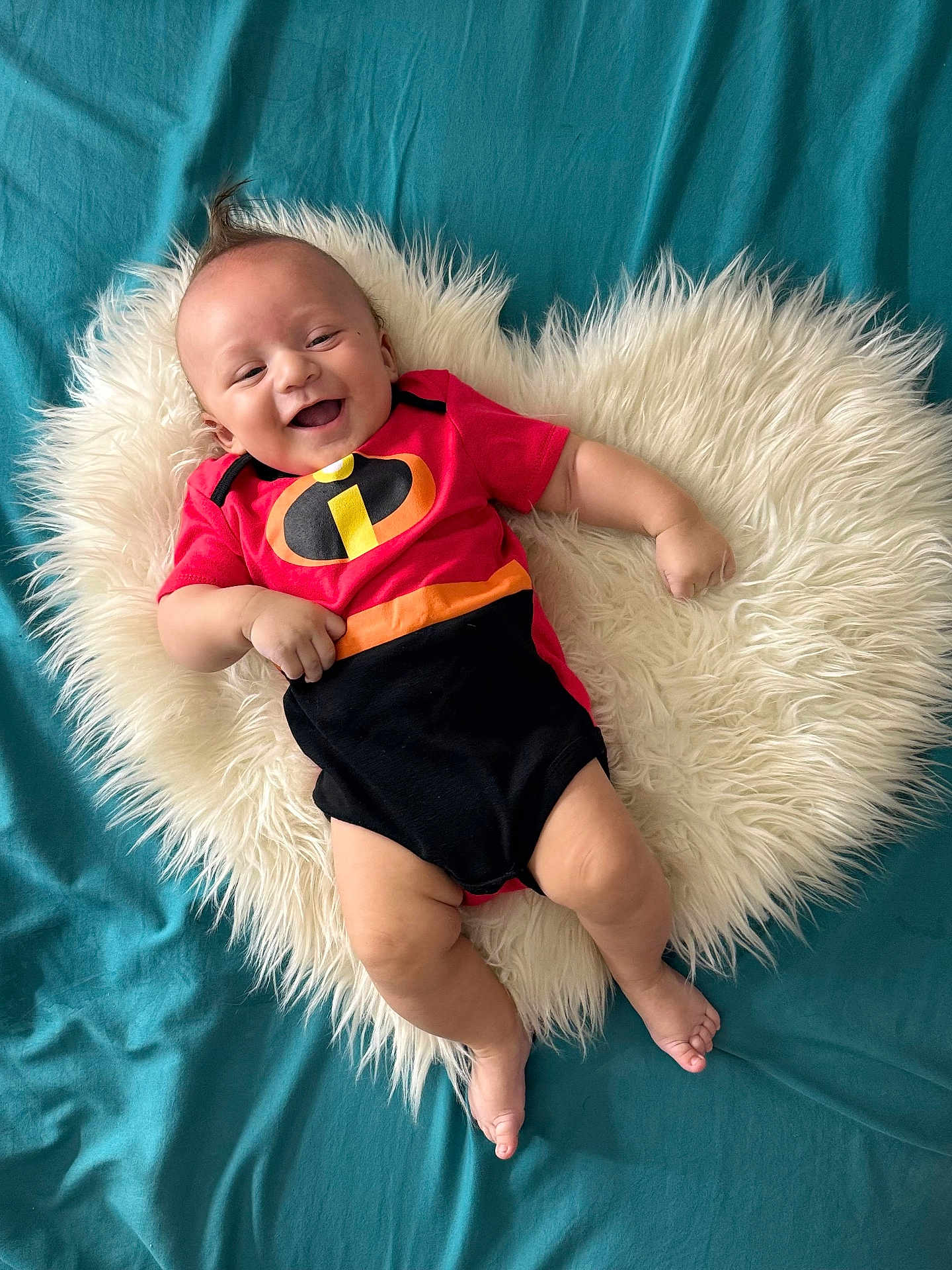 Ramón Jr is registered to the contest to win money with this photo: baby, smiling, infant, red_clothing, black_clothing, incredibles_logo, fluffy_rug, heart_shape, blue_background, lying_down, happy, cute, child, portrait, indoors, skin, bare_feet, short_sleeve, infant_fashion, baby_pose