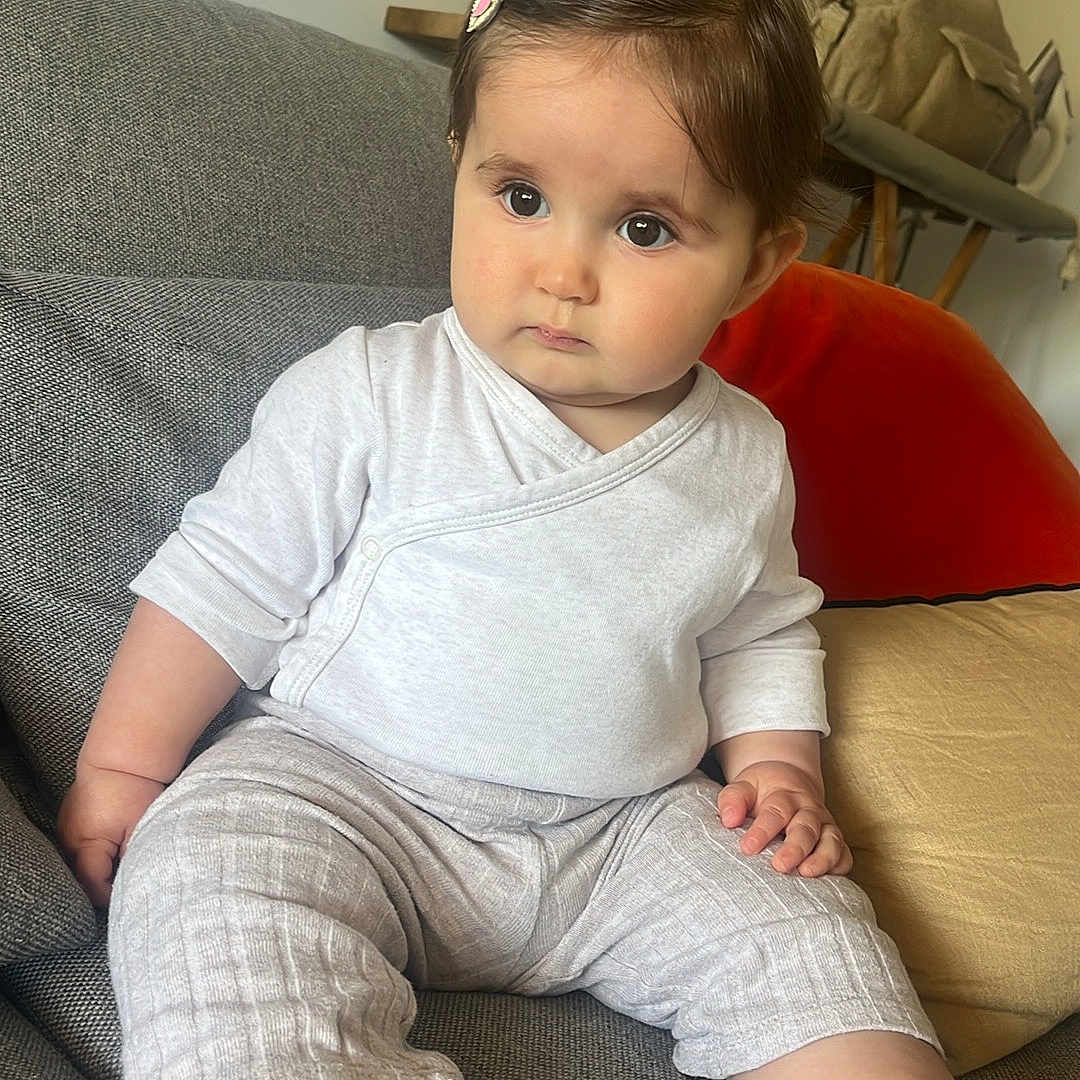Lison a rejoint le concours — aidez-le/la à gagner de superbes lots ! baby, child, infant, sitting, couch, gray_couch, toy, yellow_toy, hair_clip, clothing, pants, long_sleeve_shirt, foot, face, eyes, indoor, home, decor, painting, plant