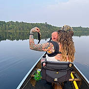 Rosey is registered to the contest to win money with this photo: adult, baby, can_holder, canoe, crying, drink_can, fish, forest, hat, lake, long_hair, nature, outdoor, paddle, person, reflection, seat, sky, tattoo, water