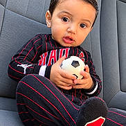 Ali is registered to the contest to win money with this photo: child, toddler, boy, portrait, sitting, car, car_seat, soccer_ball, sports, soccer, pajamas, striped_clothing, black_clothing, red_accents, short_hair, face, eyes, indoors, vehicle, happy