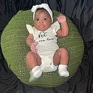Dahlani is registered to the contest to win money with this photo: baby, infant, headband, white_clothing, socks, green_cushion, knitted, raised_arm, black_background, cute, child, portrait, indoor, sitting, looking_at_camera, one_person, closeup, soft_lighting, expression, newborn