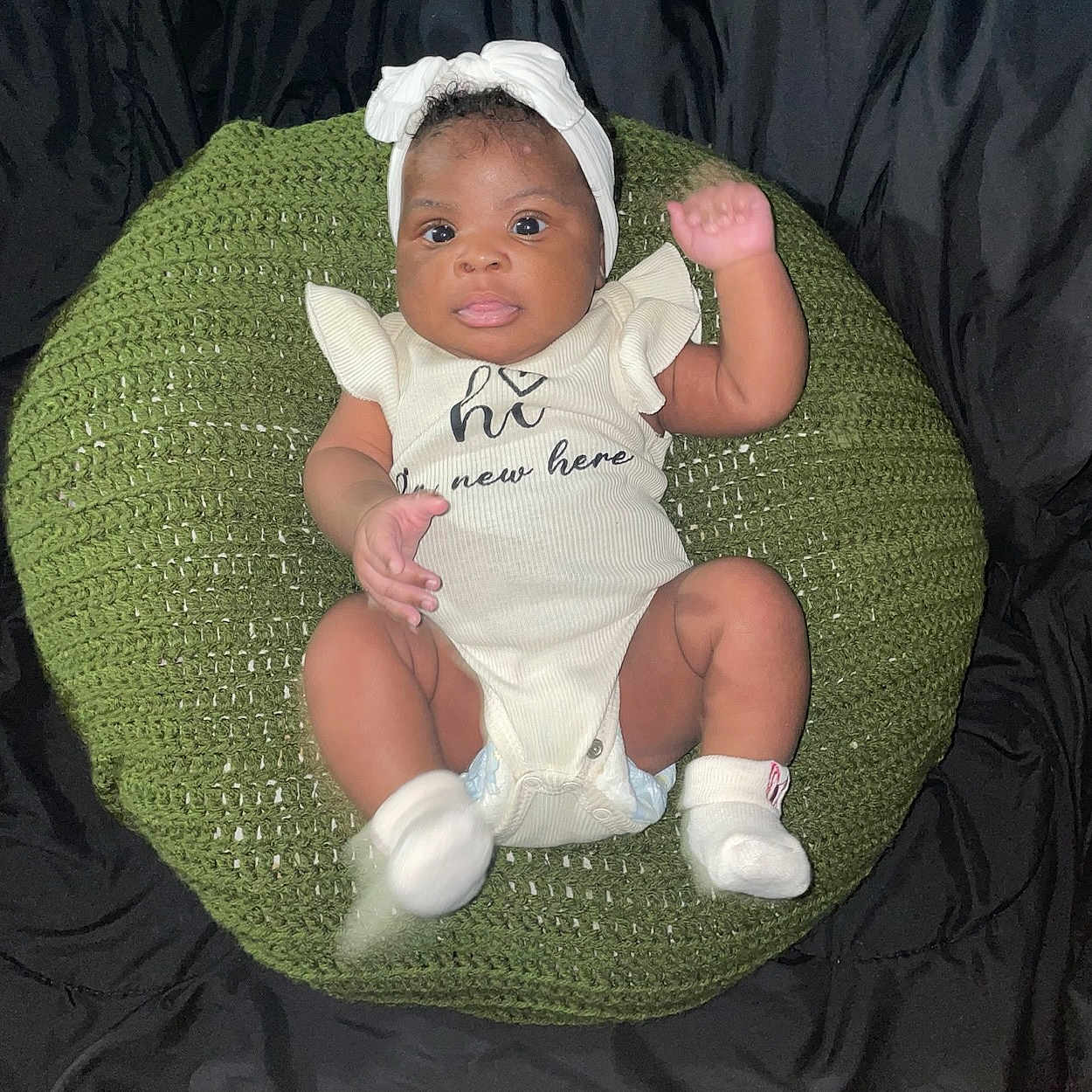 Dahlani is registered to the contest to win money with this photo: baby, infant, headband, white_clothing, socks, green_cushion, knitted, raised_arm, black_background, cute, child, portrait, indoor, sitting, looking_at_camera, one_person, closeup, soft_lighting, expression, newborn