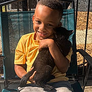 Gia joined the competition — help win amazing prizes! boy, child, cat, black_cat, smiling, hugging, outdoor, chair, yellow_shirt, gray_pants, portrait, sunlight, screened_porch, happy, pet, young, casual_clothing, resting, daytime, digital_clock_overlay