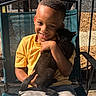 boy, child, cat, black_cat, smiling, hugging, outdoor, chair, yellow_shirt, gray_pants, portrait, sunlight, screened_porch, happy, pet, young, casual_clothing, resting, daytime, digital_clock_overlay
