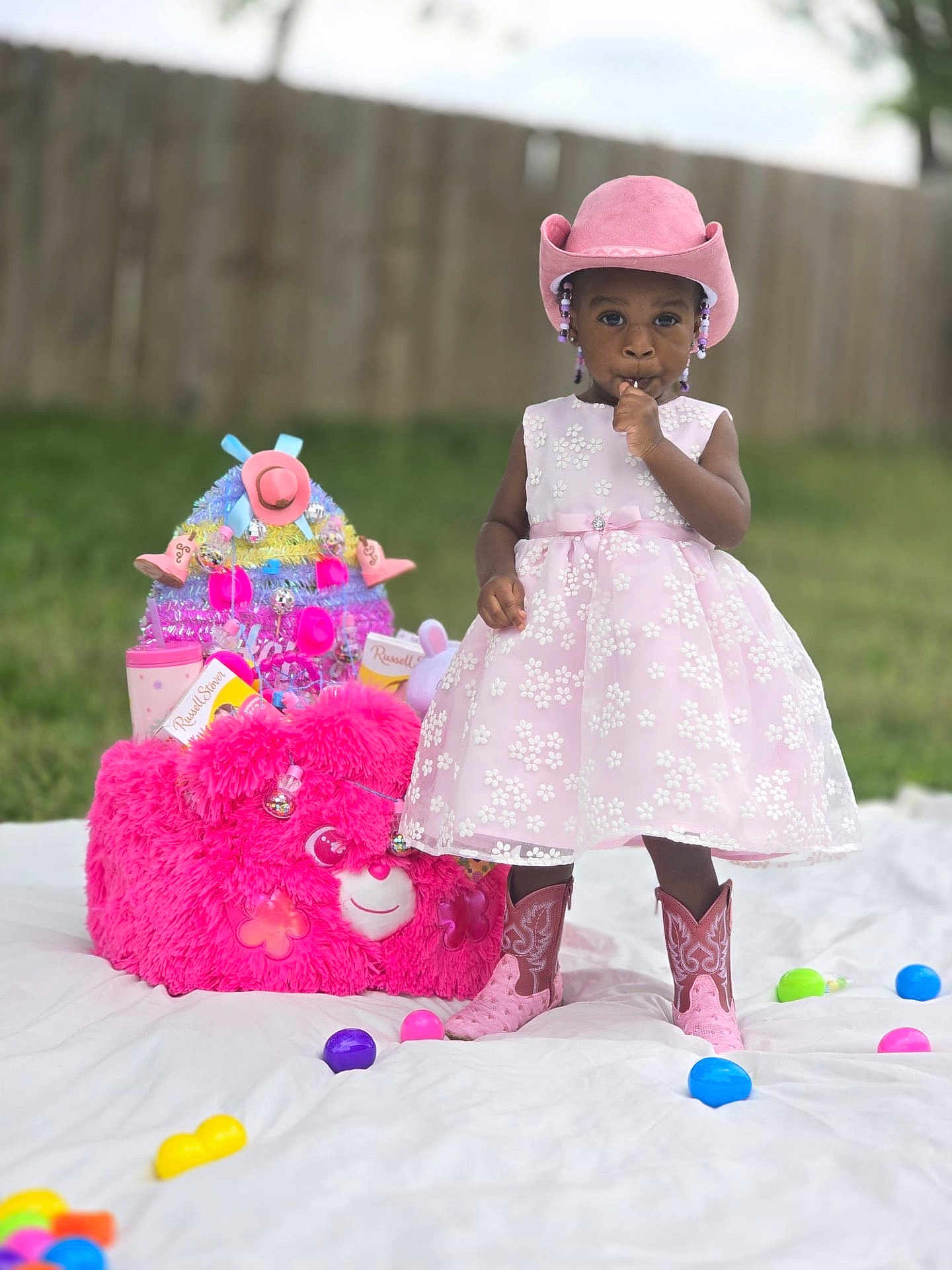 J Zelle joined the competition — help win amazing prizes! toddler, child, pink_dress, cowboy_hat, cowboy_boots, plush_toy, pink_bear, outdoor, grass, wooden_fence, colorful_eggs, toy_basket, white_blanket, beads, ribbons, sucking_thumb, standing, cute, playful, portrait