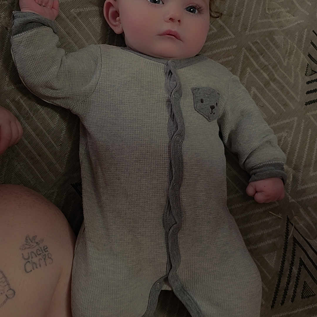 Caroline is registered to the contest to win money with this photo: baby, infant, onesie, clothing, face, human, tattoo, arm, blanket, pattern, lying_down, cute, skin, expression, child, indoors, portrait, young, person, sleepwear