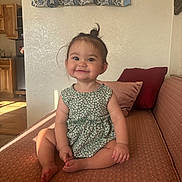 Cadence joined the competition — help win amazing prizes! toddler, child, smile, sitting, couch, living_room, curtain, patterned_dress, topknot, happy, indoors, wooden_cabinet, pillow, barefoot, face, person, home, sunlight, furniture, cozy