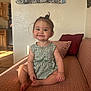 toddler, child, smile, sitting, couch, living_room, curtain, patterned_dress, topknot, happy, indoors, wooden_cabinet, pillow, barefoot, face, person, home, sunlight, furniture, cozy