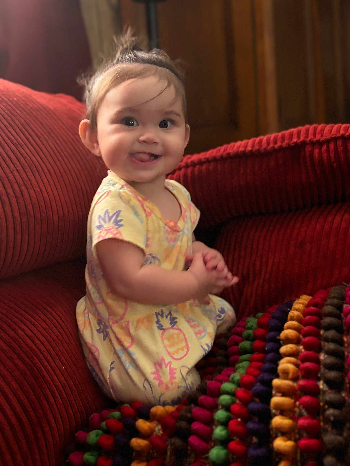 Cadence is registered to the contest to win money with this photo: baby, child, smiling, tongue_out, yellow_dress, pineapple_print, red_couch, blanket, colorful, indoor, portrait, happy, cute, playful, seated, headband, soft_lighting, furniture, young_child, person
