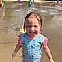 child, colorful_clothing, concrete, daylight, fun, girl, happy, kid, outdoor, park, play, recreation, smile, splash_pad, summer, sunny, swimsuit, water, water_spray, wet_hair