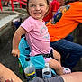 blue_chair, bottle, casual, child, crowd, daytime, event, fun, happy, outdoor, people, pink_shirt, portrait, shorts, sitting, smiling, stadium, summer, tattoo, toddler