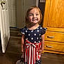 barefoot, blanket, bow, child, clothing, cute, door, dress, floor, girl, happy, home, indoor, person, portrait, smile, standing, stars, stripes, wooden_furniture