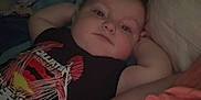 Anthony joined the competition — help win amazing prizes! baby, infant, child, person, face, arms, shirt, metallica, text_on_clothing, blanket, pillow, lying_down, indoor, relaxed, looking_at_camera, dim_light, casual, resting, bedroom, cozy