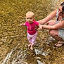 baby, child, woman, water, creek, rocks, nature, outdoor, barefoot, supporting, smiling, pink_clothing, person, shallow_water, riverbed, grass, summer, daylight, holding_hands, happy