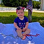 Finnley is registered to the contest to win money with this photo: baby, child, outdoor, grass, tree, flag, american_flag, headband, blue_clothing, beads, blanket, sunlight, summer, patriotic, smile, sitting, cute, infant, nature, holiday