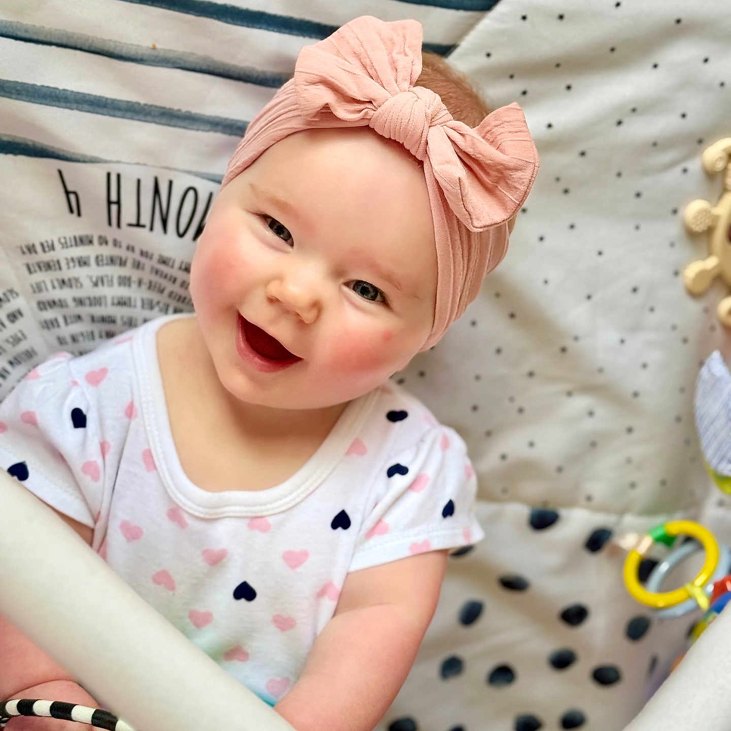 Emilia is registered to the contest to win money with this photo: baby, child, smiling, headband, pink, toy, playmat, polka_dot, striped, cheeks, clothing, infant, happy, cute, indoors, person, portrait, holding, colorful, closeup