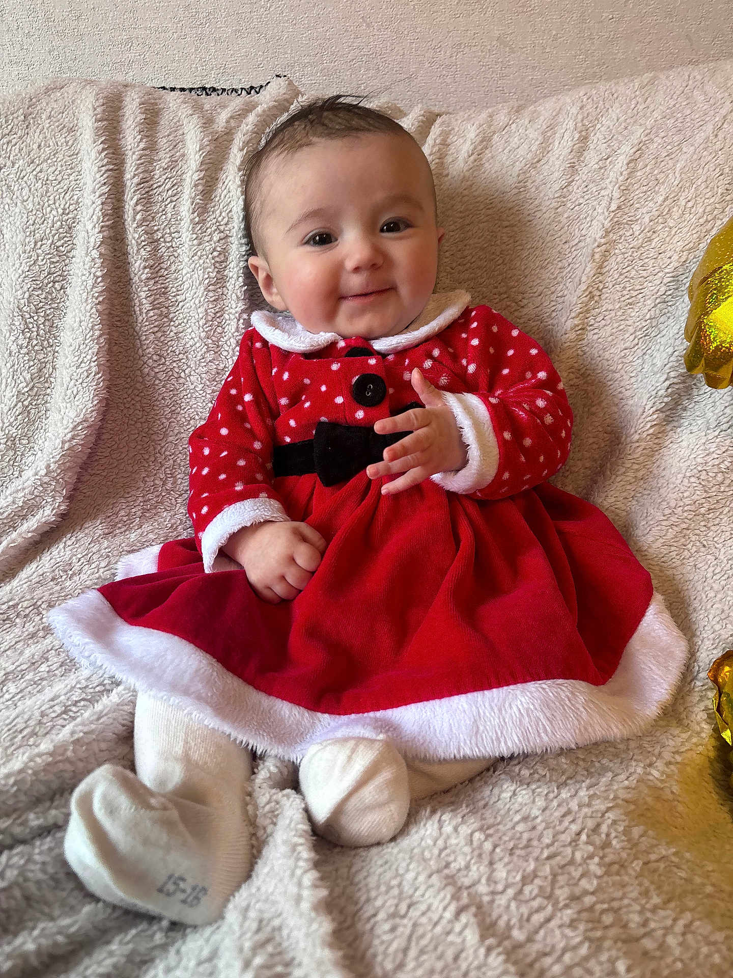 Inaya a rejoint le concours — aidez-le/la à gagner de superbes lots ! baby, child, smiling, red_dress, white_trim, sitting, blanket, indoors, cute, holiday_outfit, festive, cozy, portrait, happy, infant, clothing, soft_texture, face, person, warm
