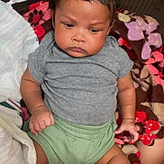 Messiah is registered to the contest to win money with this photo: baby, infant, child, blanket, clothing, onesie, green_shorts, curly_hair, face, expression, indoor, furniture, couch, patterned_blanket, skin, hand, foot, portrait, serious, sitting