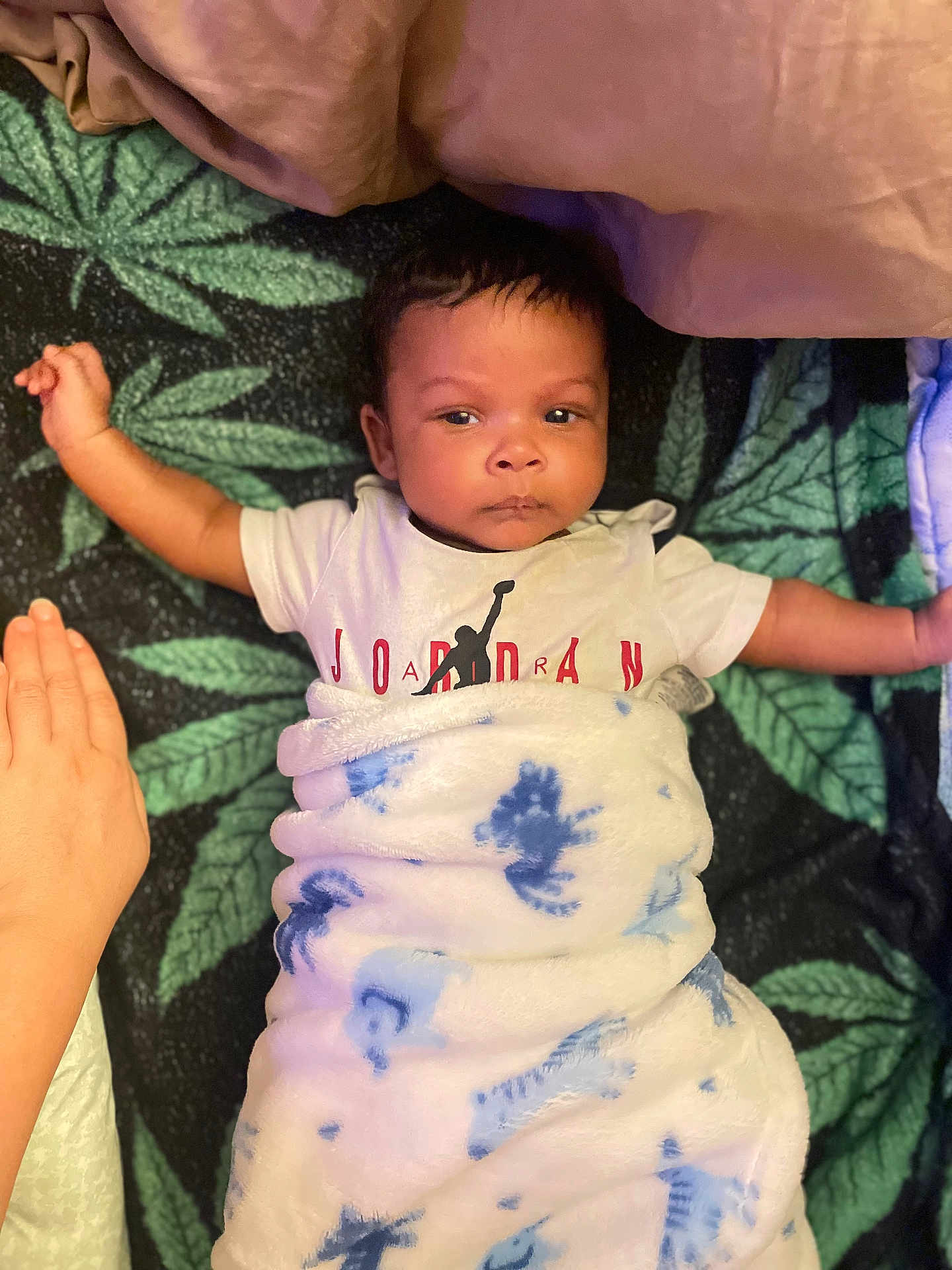 Messiah is registered to the contest to win money with this photo: baby, infant, blanket, tshirt, person, face, hand, lying_down, leaf_pattern, soft_texture, indoor, child, wrapped, clothing, cute, young, portrait, resting, peaceful, comfort