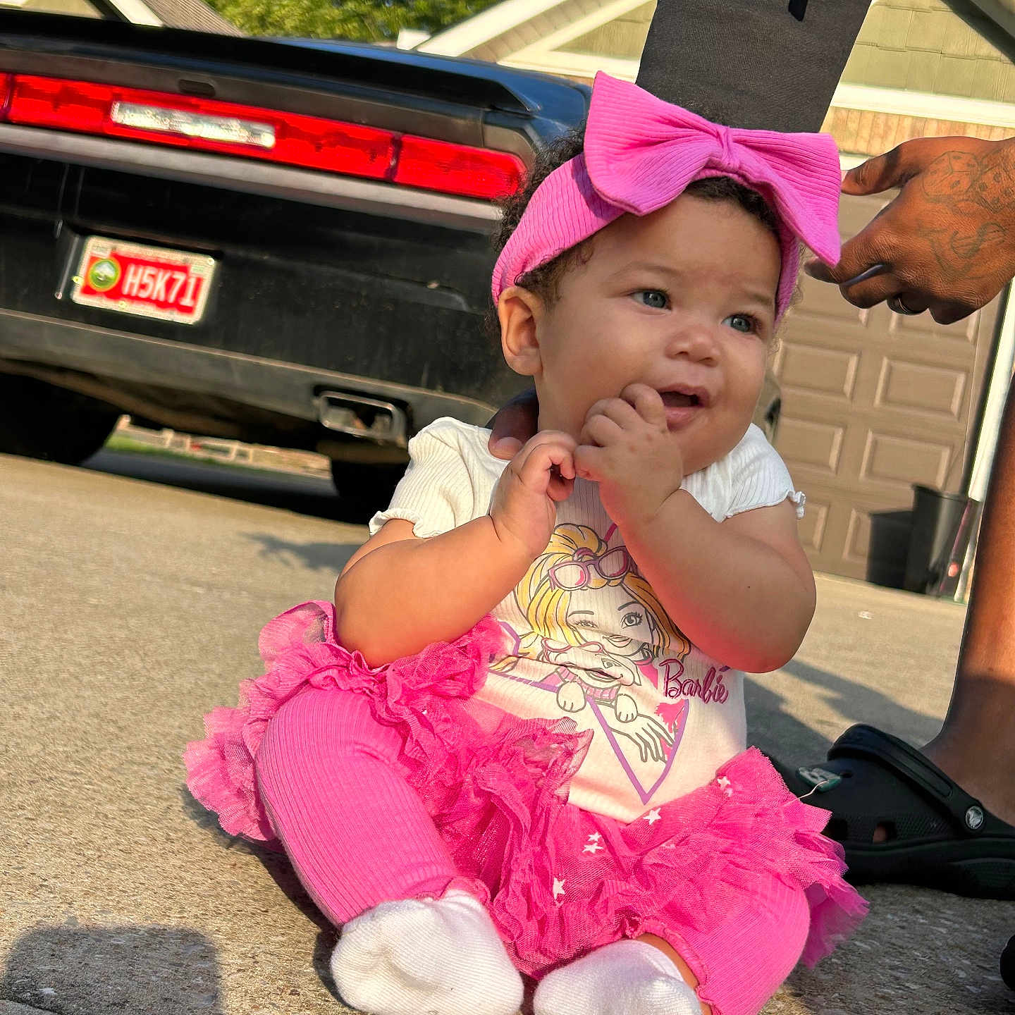 Jeylani is registered to the contest to win money with this photo: adult_hand, baby, black_sandal, car, child, cute, driveway, footwear, house, outdoor, person, pink_bow, pink_leggings, pink_skirt, portrait, smiling, sunlight, tattoo, tshirt, white_socks