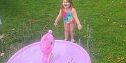 Skylar is registered to the contest to win money with this photo: child, girl, swimsuit, water, sprinkler, inflatable, care_bears, grass, lawn, outdoor, summer, play, fun, smiling, happy, tree, shed, car, toy, nature