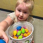 Skylar is registered to the contest to win money with this photo: child, toddler, bucket, plastic_eggs, easter_eggs, indoor, floor, wall, yellow_wall, tile_floor, person, curly_hair, pony_tail, hand, colorful, toy, offering, expression, young_child, clothing