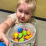 child, toddler, bucket, plastic_eggs, easter_eggs, indoor, floor, wall, yellow_wall, tile_floor, person, curly_hair, pony_tail, hand, colorful, toy, offering, expression, young_child, clothing