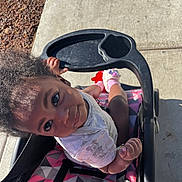 Karizma is registered to the contest to win money with this photo: child, toddler, stroller, sidewalk, tray_table, curly_hair, baby_clothing, socks, pink, seat, looking_up, hand, outdoor, sunlight, pavement, patterned_seat, expression, toy, portrait, happy
