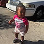 Karizma is registered to the contest to win money with this photo: child, toddler, pink_shirt, shorts, sneakers, socks, sidewalk, concrete, white_car, parked_car, head, face, curious_expression, shadow, sunlight, parking_lot, urban, curb, hair_bun, standing