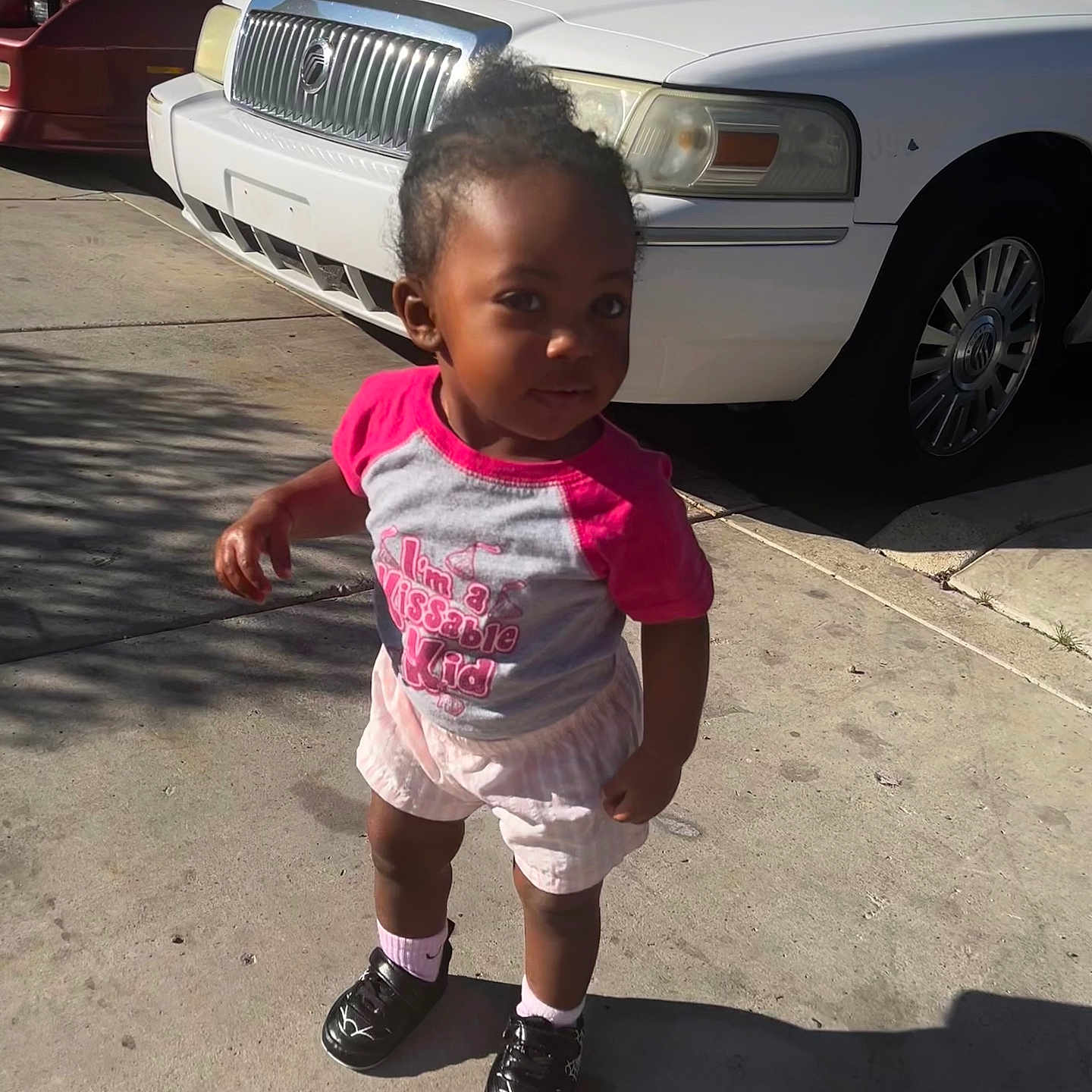 Karizma is registered to the contest to win money with this photo: child, concrete, curb, curious_expression, face, hair_bun, head, parked_car, parking_lot, pink_shirt, shadow, shorts, sidewalk, sneakers, socks, standing, sunlight, toddler, urban, white_car