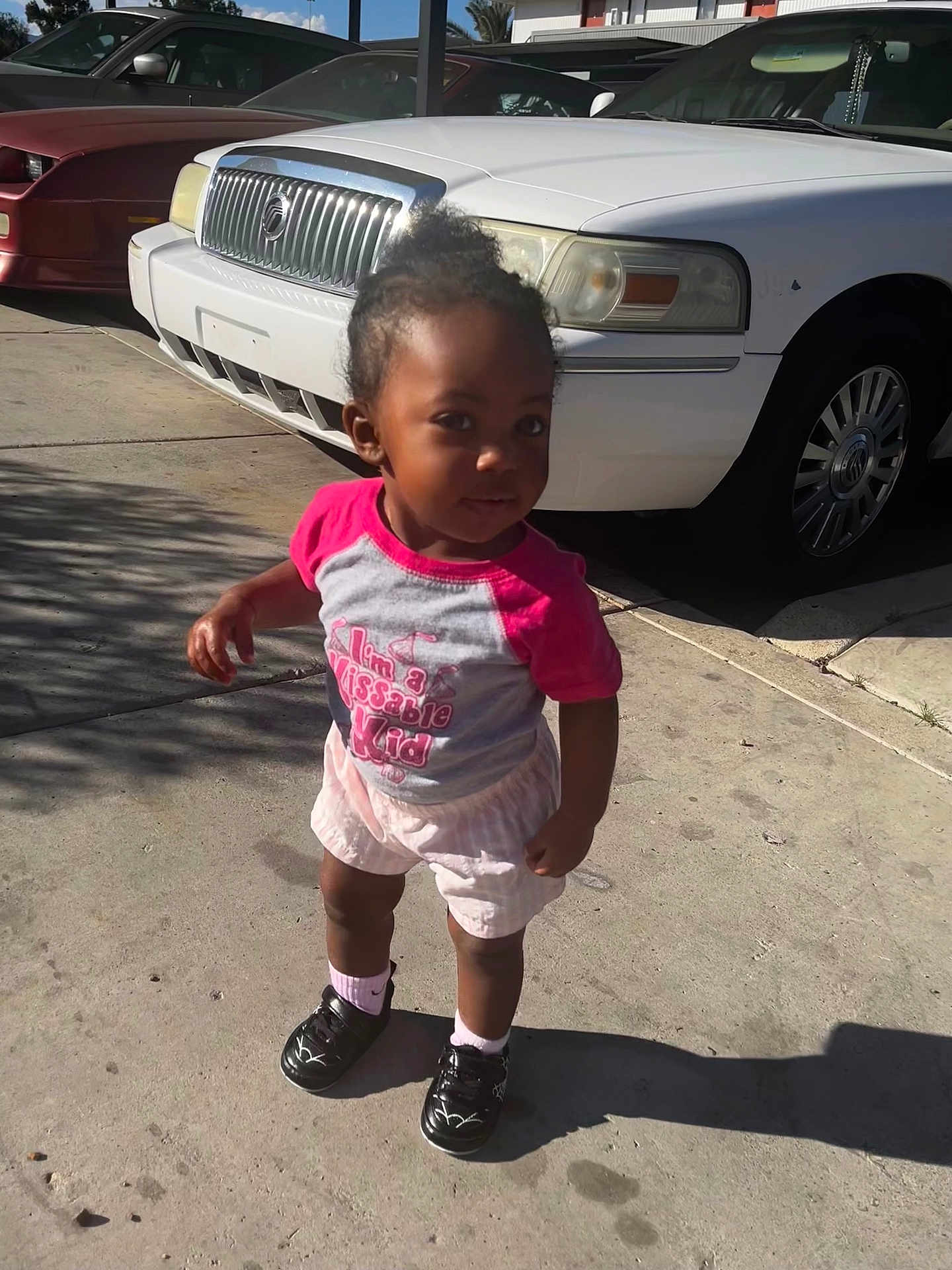 Karizma is registered to the contest to win money with this photo: child, toddler, pink_shirt, shorts, sneakers, socks, sidewalk, concrete, white_car, parked_car, head, face, curious_expression, shadow, sunlight, parking_lot, urban, curb, hair_bun, standing