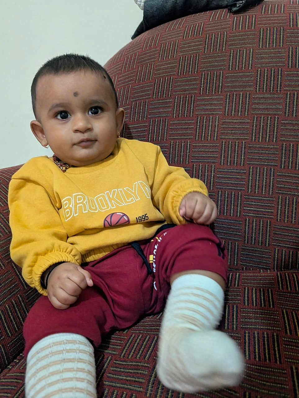 Sharavin is registered to the contest to win money with this photo: baby, child, yellow_sweatshirt, maroon_pants, striped_socks, couch, patterned_fabric, indoors, portrait, cute, sitting, small_ears, short_hair, expression, comfortable, furniture, clothing, person, young_child, casual