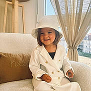 Zoé a rejoint le concours — aidez-le/la à gagner de superbes lots ! child, toddler, smiling, white_suit, bucket_hat, indoor, armchair, cushion, curtains, window, natural_light, lamp, wooden_lamp, happy, portrait, cozy, home, person, fashion, cute