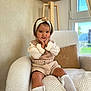 armchair, child, cozy, cushion, happy, headband, home, indoor, knitwear, lamp, long_sleeve_shirt, natural_light, plush, shoes, shorts, sitting, smile, sock, toddler, window