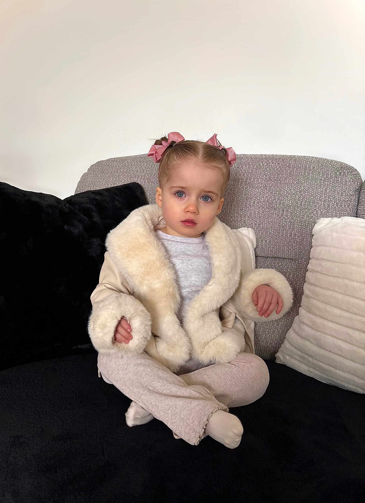 Evelyn joined the competition — help win amazing prizes! toddler, child, girl, pink_bows, fur_jacket, beige_pants, socks, couch, pillows, indoor, portrait, cross_legged, blue_eyes, curious_expression, furniture, soft_lighting, casual_clothing, seated, young_child, home