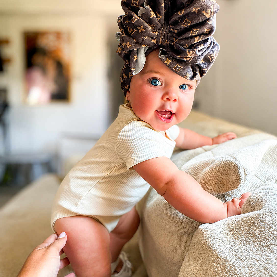 Haddie joined the competition — help win amazing prizes! baby, child, blue_eyes, headwrap, onesie, couch, hand, indoor, smiling, happy, infant, person, skin, furniture, living_room, curious, support, cute, closeup, portrait