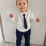 black_shoes, child, curious, cute, indoor, navy_pants, pants, person, plain_background, shirt, short_hair, smile, sneakers, standing, tie_print, tile_floor, toddler, vest_print, white_shirt, young_child