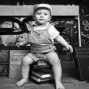 Alessio participe au concours pour gagner de l'argent avec cette photo : baby, barefoot, black_and_white, books, cap, chalkboard, child, cute, desk, floor, indoor, overalls, papers, person, portrait, sitting, smiling, stack, vintage, wooden_floor