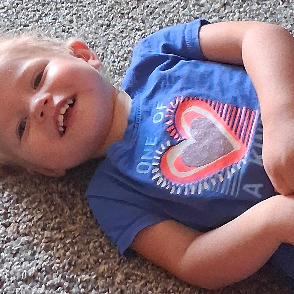 Nataila joined the competition — help win amazing prizes! blond_hair, blue_shirt, carpet, casual_clothing, child, childhood, cute, fun, happy, heart_print, indoor, lying_down, person, playing, portrait, relaxed, small_hands, smiling, toddler, young_child