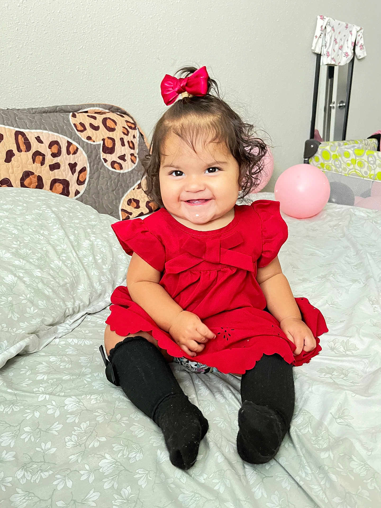 Khloe Mahiara is registered to the contest to win money with this photo: baby, child, smiling, red_dress, bow, black_socks, bed, pillow, balloon, indoor, cute, happy, portrait, sitting, curly_hair, drool, toddler, wall, blanket, infant