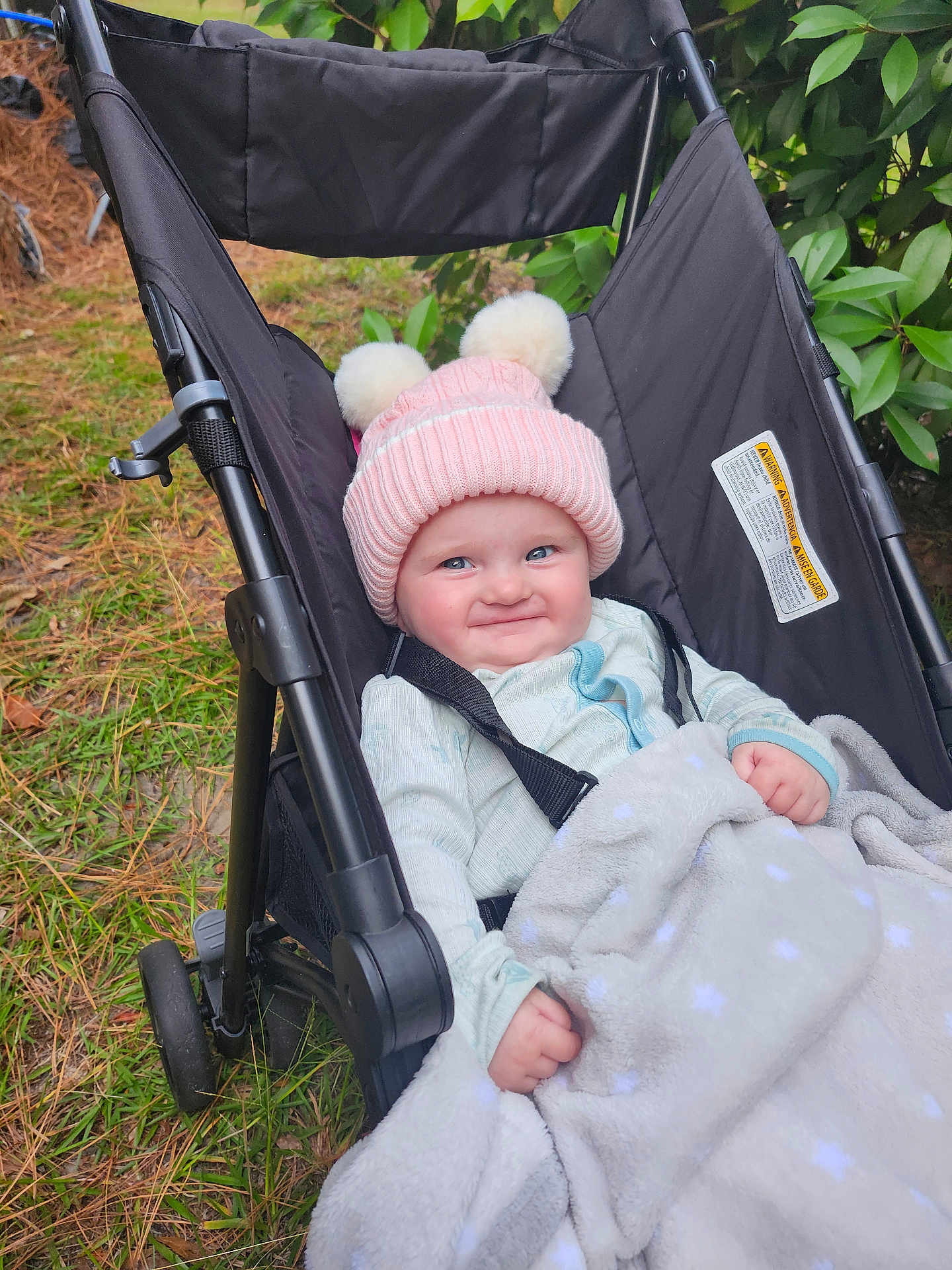 Aurora joined the competition — help win amazing prizes! baby, black_stroller, blanket, child, cozy, cute, face, grass, greenery, happy, infant, nature, outdoor, pajamas, person, pink_hat, pom_poms, portrait, smile, stroller